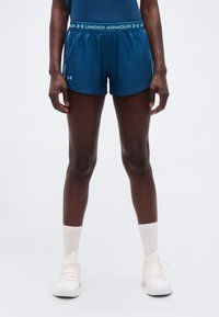 Άτομο που στέκεται φορώντας μπλε αθλητικά σορτς Under Armour, λευκές κάλτσες πλήρους μήκους και λευκά αθλητικά παπούτσια, μπροστά σε απλό φόντο.