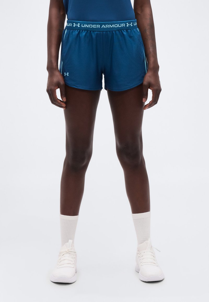 Άτομο που στέκεται φορώντας μπλε αθλητικά σορτς Under Armour, λευκές κάλτσες πλήρους μήκους και λευκά αθλητικά παπούτσια, μπροστά σε απλό φόντο.