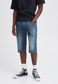 Shorts en denim bleu avec une coupe droite, ourlets retournés et décoloration subtile. Associés à des baskets montantes noires et des chaussettes blanches mi-mollet.