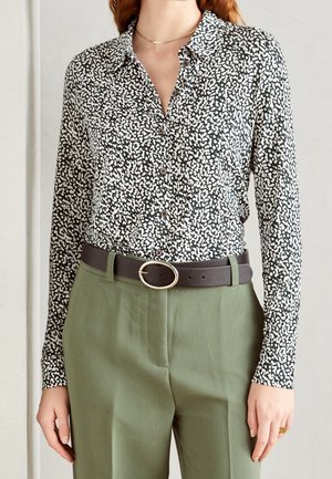 Kvinde iført en sort og hvid skjorte med abstrakt print, knappet op og stoppet ind i højtaljede olivengrønne bukser med sort bælte.