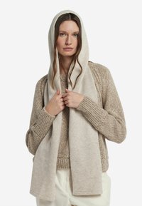 Beżowy szalik z miękkiego materiału, drapowany na ramionach w oversize'owym stylu. Dzianinowy sweter o fakturze w jaśniejszym odcieniu dopełnia wygląd.