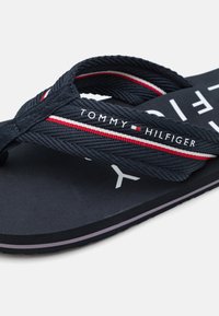 Încălțăminte tip flip-flop marin cu curele împletite, evidențiate de accente roșii și albe, talpă texturată și logo de marcă pe talpă. Talpă exterioară din cauciuc plat.