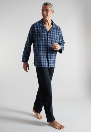 Mann mit grauen Haaren, der lächelt und barfuß läuft, trägt ein blau-schwarz kariertes Hemd mit Knöpfen und schwarze Hose vor einem einfarbigen Hintergrund.