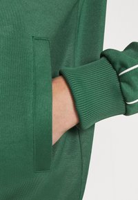 Sweat à capuche vert avec des poignets côtes, une poche latérale et un tissu au toucher lisse. Présente un design simple sans motifs ni accents visibles.