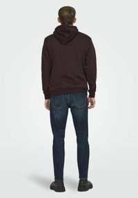 Sudadera con capucha color marrón oscuro, con bolsillo tipo canguro, dobladillo y puños de canalé, combinada con jeans ajustados de color azul oscuro y zapatos negros.