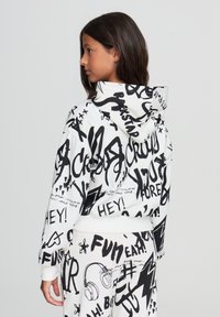 Felpa bianca con testo e elementi grafici in stile graffiti neri. Presenta una vestibilità rilassata e un cappuccio con coulisse. Abbinata a pantaloni coordinati.