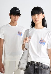 White cotton t-shirts feature a blue and orange graphic logo. A gray crossbody bag is held by one model. Simple, unadorned design.