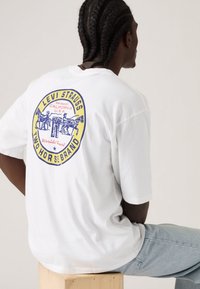 Λευκό βαμβακερό μπλουζάκι με πολύχρωμο κυκλικό γραφικό Levi Strauss στην πλάτη, με μπλε και κίτρινες αποχρώσεις και άνετη εφαρμογή.