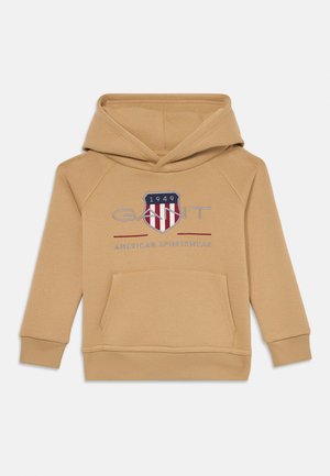 Tan hoodie with a kangaroo pocket, featuring a shield logo in blue, red, and white. Ribbed cuffs and hem, made from soft fabric.