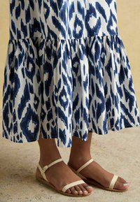 Zilā un baltā rakstaina maksi kleita ar viļņotu apakšējo malu, apvienota ar brūniem plakaniem sandālēm, kuru dizainā ir divi šauri siksniņas un sprādze.