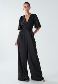 Sievietei mugurā ir melna plaša kombinezona uzvalks ar dziļu V-veida kakla izgriezumu un īsām puff piedurknēm, apskaņots ar zelta auskariem un rokassprādzēm.