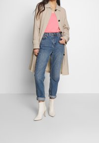 Beige trenchcoat päällä vaaleanpunainen v-kaula-aukkoinen paita, yhdistettyinä käännettyihin sinisiin farkkuihin ja valkoisiin nilkkabootseihin. Silkkinen kangas, jossa on ryhdikäs istuvuus.
