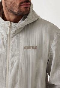 Svetlo šedá rebrovaná bunda s kapucňou, s predným zipsom a logom "GUESS" v kontrastnej béžovej farbe. Hladká textúra látky.