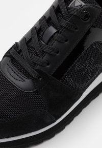 Gros plan sur une basket noire montrant des matériaux en mesh, cuir et daim, avec des lacets noirs et une semelle à rayures blanches sur un fond clair.