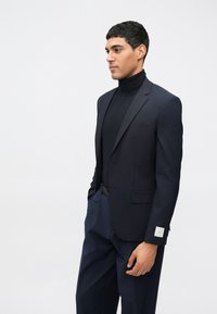 Blazer azul-marinho com um corte ajustado, fecho de botão único e um lenço de bolso. Usado sobre um suéter preto de gola alta, combinado com calças a condizer.