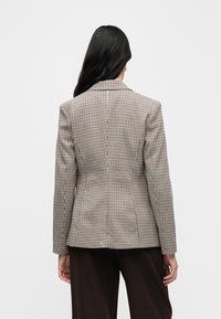 Femme aux longs cheveux foncés portant un blazer beige et noir à carreaux et un pantalon foncé, vue de dos sur un fond uni.