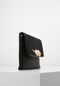 Pochette en cuir noir avec un rabat en daim orné d'un embellissement métallique doré en forme de feuille, posée sur une surface blanche.