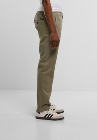 Pantalones caqui de corte recto, con cintura elástica, bolsillos laterales y una etiqueta. Combinados con zapatillas blancas y negras con suela texturizada.