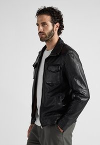 Zwarte leren jas met een ritsluiting, twee voorzakken en een klassieke kraag. Draagt over een grijs shirt en gecombineerd met een grijze broek.