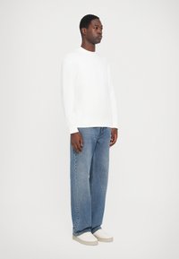 Sudadera blanca con cuello redondo, combinada con jeans de corte ancho en azul claro y zapatillas blancas. Textura suave y diseño sencillo.