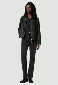 Man with long dreadlocks wearing a black leather jacket, black button-up shirt, black jeans, and black boots, standing with one hand in pocket.