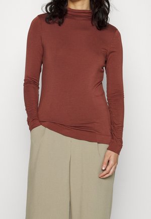 Kobieta ubrana w dopasowany, długi rękaw, rdzawy golf i luźne beżowe spodnie, z rękami w kieszeniach.