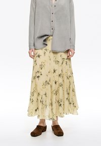 Lange gele bloemenrok met zwarte patronen, gelaagd ontwerp, gecombineerd met een losse grijze blouse met knopen en bruine suède instapschoenen.