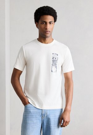 Bărbat cu coafură afro, purtând un tricou alb cu imprimeu grafic și blugi albastru deschis, stând cu o mână în buzunar, pe fundalul unui perete simplu.