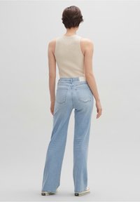 Persona con cabello corto castaño, vistiendo una blusa sin mangas de punto beis, jeans de tiro alto de pierna ancha en azul claro y zapatillas blancas, de espaldas.