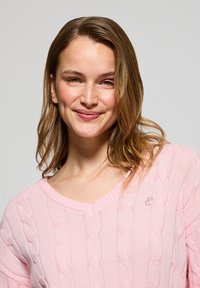 Ljusrosa kabelstickad tröja med V-ringning, med en subtil logodetalj. Mjuk textur och avslappnad passform med långa ärmar.