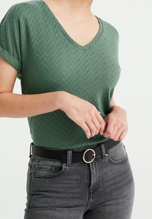 Vrouw draagt een groen gestructureerd V-hals T-shirt met korte mouwen, ingestopt in een hoog-uitgesneden zwarte spijkerbroek met een zwarte riem met een gouden ronde gesp.