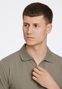Olivengrøn polo skjorte med krave, lavet af blødt stof. Modellen justerer skjortens krave med den ene hånd.