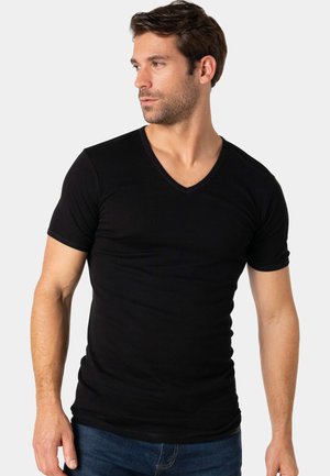 Homme aux cheveux courts et foncés portant un t-shirt noir près du corps à col en V et un jean bleu, regardant vers sa gauche sur un fond uni.