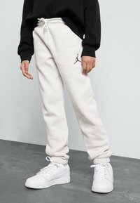 Pantalones de chándal de felpa blanca con cinturilla y puños elásticos, que cuentan con un logo Jumpman gris en el muslo izquierdo, combinados con zapatillas blancas.