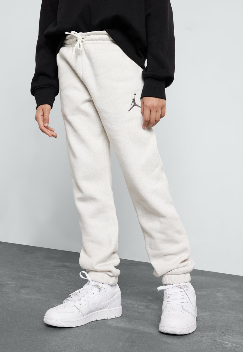 Pantalones de chándal de felpa blanca con cinturilla y puños elásticos, que cuentan con un logo Jumpman gris en el muslo izquierdo, combinados con zapatillas blancas.