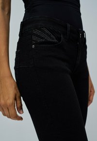 Pantalones vaqueros de mezclilla negra con un diseño ajustado, textura suave y un patrón decorativo de piedras de strass en el interior del bolsillo delantero.