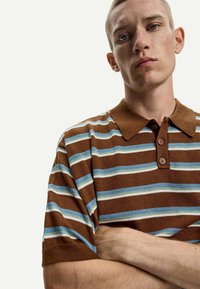 Polo shirt lavorato a maglia di colore marrone con righe blu e bianche, dotato di colletto e patta con tre bottoni. Texture morbida e maniche corte.