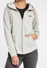 Sweat à capuche gris clair zippé avec un motif à pois, capuche ajustable et poches avant. Comprend des cordons de serrage contrastants et des poignets côtelés.
