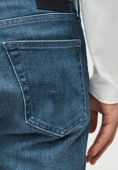 Πίσω τσέπη από μπλε τζιν με ραφές, φορεμένη από άτομο με λευκό μακρυμάνικο πουκάμισο, το χέρι ακουμπά κοντά στην τσέπη.