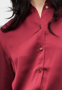 Γυναίκα που φοράει μπλούζα από σκούρο κόκκινο μετάξι με κουμπιά σε χρυσούς τόνους, εμφανίζοντας λεπτομέρειες στο γιακά και την περιοχή του πάνω στήθους.