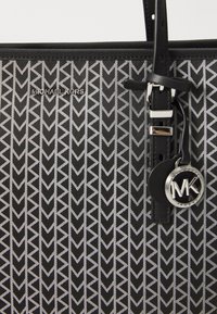 Black tote bag featuring a silver geometric pattern of arrows, with a logo accent and leather strap detail. Smooth texture, structured shape.