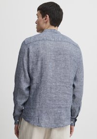 Herreshirt i teksturert blått stoff, med krage med knapper, lange ermer og avrundet hem. Bakvisningen viser subtile mønstre.