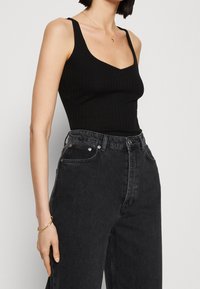 Černý žebrovaný top bez rukávů s kulatým výstřihem, který je kombinovaný s černými džíny s vysokým pasem. Má uvolněný střih a zlatý náramek.