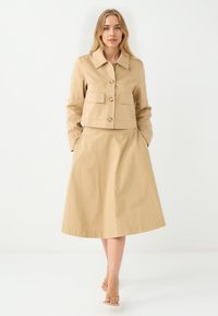 Beige cropped Jacke mit großen Fronttaschen, kombiniert mit einem passenden A-Linien-Rock. Beide haben eine glatte Textur und minimale Details.