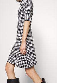 Robe à motif pied-de-poule noir et blanc avec une jupe évasée, des manches courtes et un corsage ajusté, fabriquée dans un tissu léger.