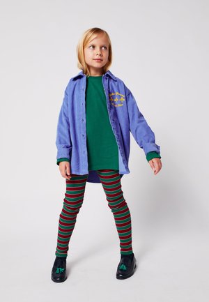 Hellblaues übergroßes Button-Down-Hemd über einem grünen T-Shirt, kombiniert mit rot-grün gestreiften Leggings und schwarzen Schuhen mit Quasten.