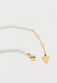 Σειρά από λευκές πέρλες με χρυσό κούμπωμα τύπου αστακού και τριγωνικό μενταγιόν με τη λέξη "GUESS". Διαθέτει προέκταση από αλυσίδα με κρίκους.