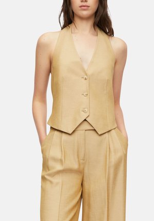 Frau trägt eine ärmellose beige maßgeschneiderte Weste mit drei Knöpfen und passende, hoch taillierte plissierte Hosen, Hände in den Taschen.