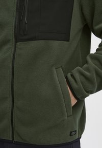 Grønn fleecejakke med en svart nylon brystlomme, glidelås, elastiske mansjetter og sidelomme med en liten merkeetikett nederst.