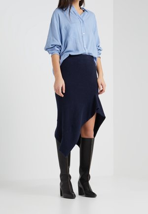 Femme portant un chemisier bleu clair à manches longues, une jupe asymétrique bleu marine et des bottes noires montantes à talons, debout devant un fond blanc.